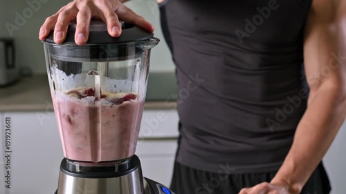 Man adds banana to blender with berries and protein powder, then starts blending in the kitchen for a healthy shake.
