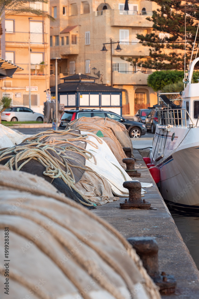 Fototapeta premium Roquetas De Mar, Spain - June 03, 2025: Folded fishing gear on waterfront by bollards against background of moored boats and coastal city buildings in warm evening light.