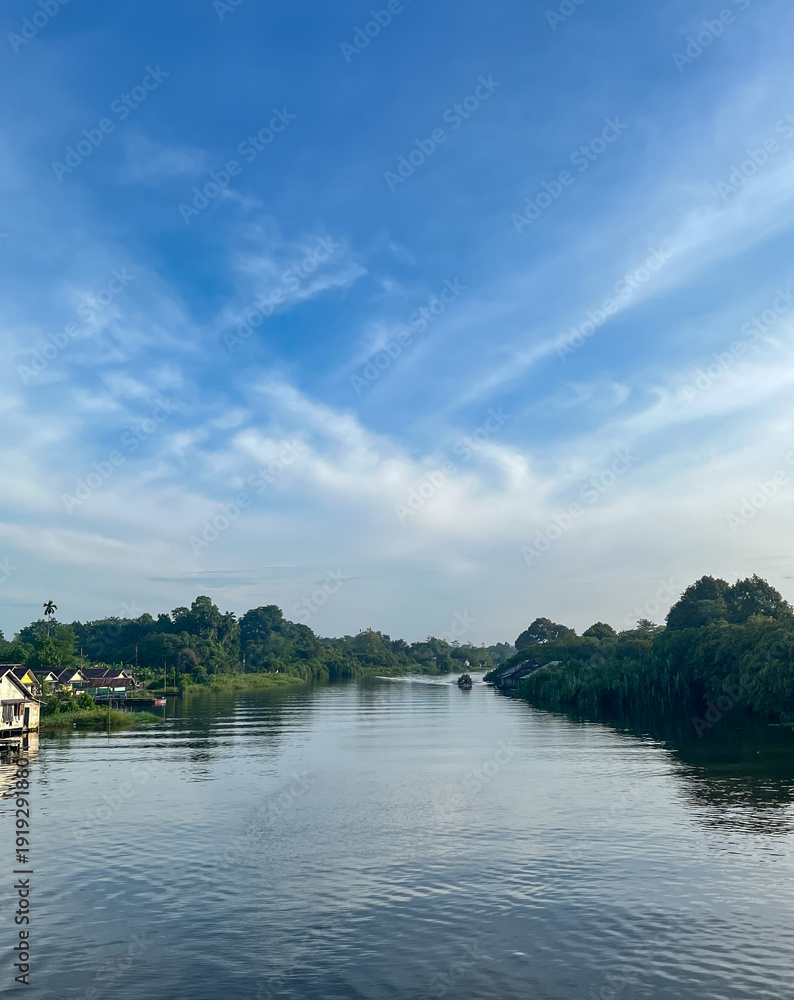 Fototapeta premium Calm river flowing between lush green banks with riverside houses under a wide blue sky with soft clouds.