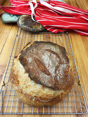 Closeup of a freshly baked loaf of crusty bread on a cooling rack, with an apron and oven gloves behind.