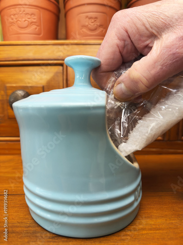 Closeup of a hand pouring sea salt flakes from a clear bag into a stoneware salt keeper (also called a salt pig).