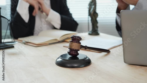 An Asian businessman and a female lawyer, both in suits, are examining documents with hammers at a wooden table near a window in an office, indicating that legal proceedings are underway.