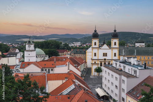 Evening cityscape of Trencín with Piarist Church of St. Francis Xavier, City Tower, red rooftops and hills under pastel sunset sky. Historic Slovak town