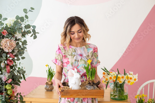 woman in floral dress holding wooden tray with spring bulbs and easter bunny decoration in pastel studio, festive portrait celebrating easter and womens day