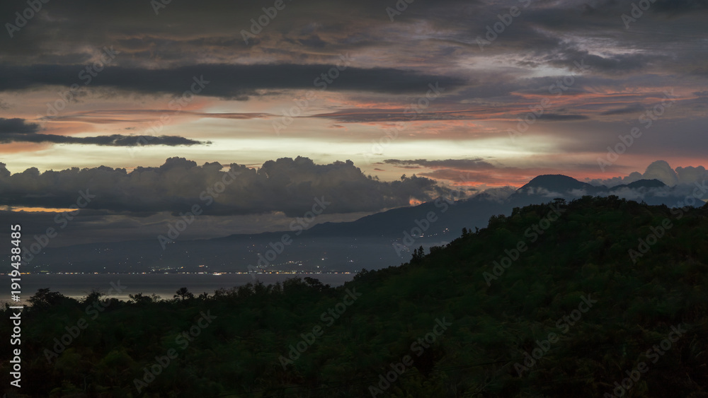 Fototapeta premium Layered clouds stretch across the evening sky above dark forested hills, with distant coastal water and scattered lights visible below. 