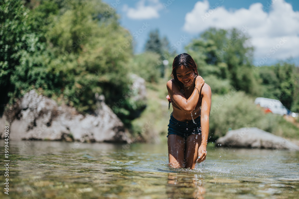 Fototapeta premium A young woman stands in clear river water, smiling as she splashes. The sunny outdoor scene shows green trees, rocks, and a relaxed summer vibe.