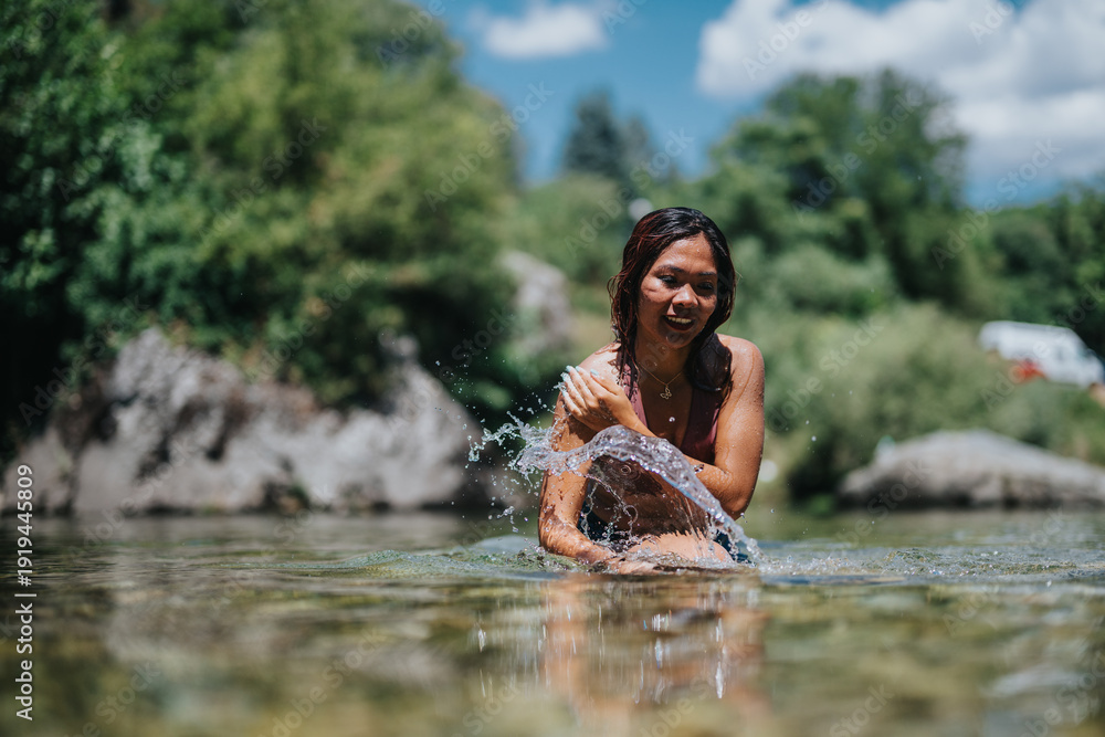 Fototapeta premium A woman smiles and splashes water while standing in a shallow riverside. Bright outdoor scene with trees, rocks, and a blue sky.