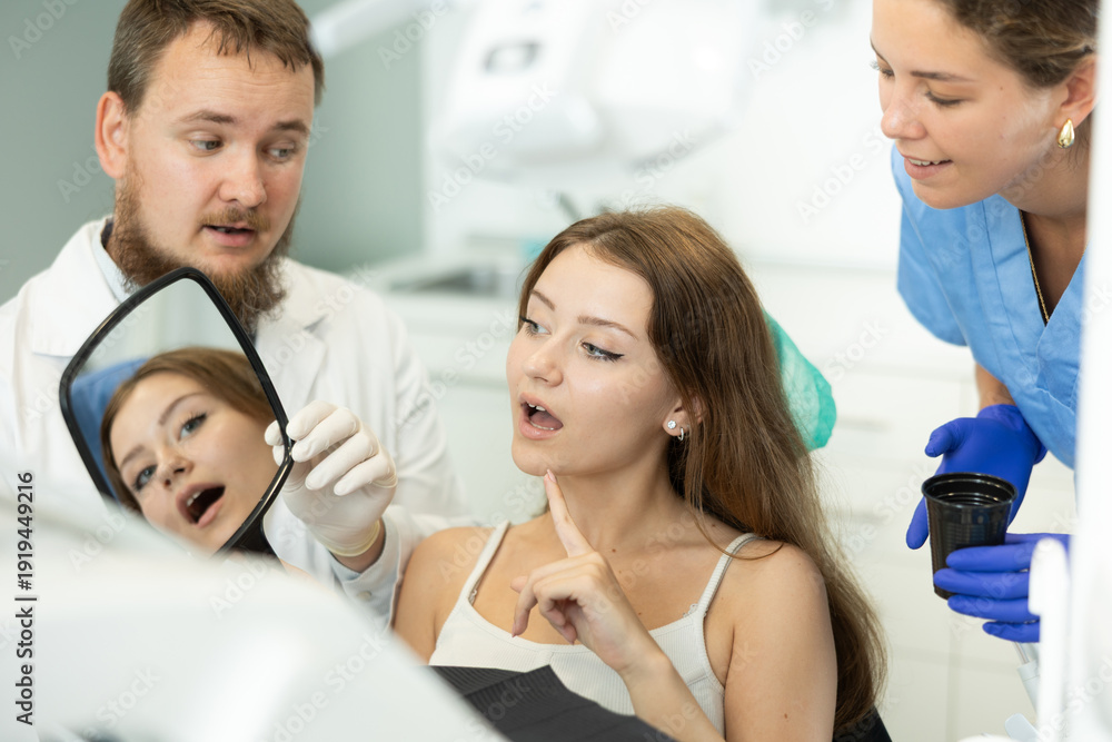 Obraz premium Interested young woman sits in a dentist chair and carefully looks at her teeth in the mirror. Male dentist with an assistant shows the client the work done