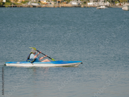 A solo kayaker drifts in a blue sit-on-top kayak on calm bay lake water, wearing a sun hat and life vest while paddling slowly and enjoying a peaceful summer outdoor adventure.
