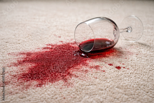 Spilled red wine on light carpet near overturned glass. Stain on textile floor at home interior, concept of household accident and cleaning problem.
