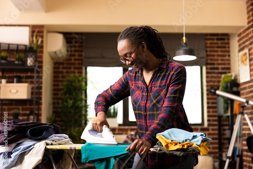 Quadro su tela Smiling black housewife using electric iron to press clean shirt, removing wrinkles after laundry