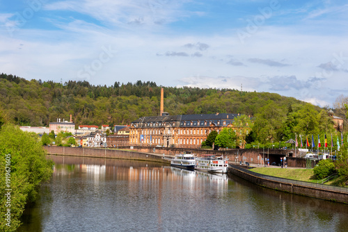Scenic view Mettlach historic architecture industrial heritage Saar river Germany forested hills calm waterfront. Scene panorama cruise boat brick buildings, chimney vibrant spring greenery blue sky