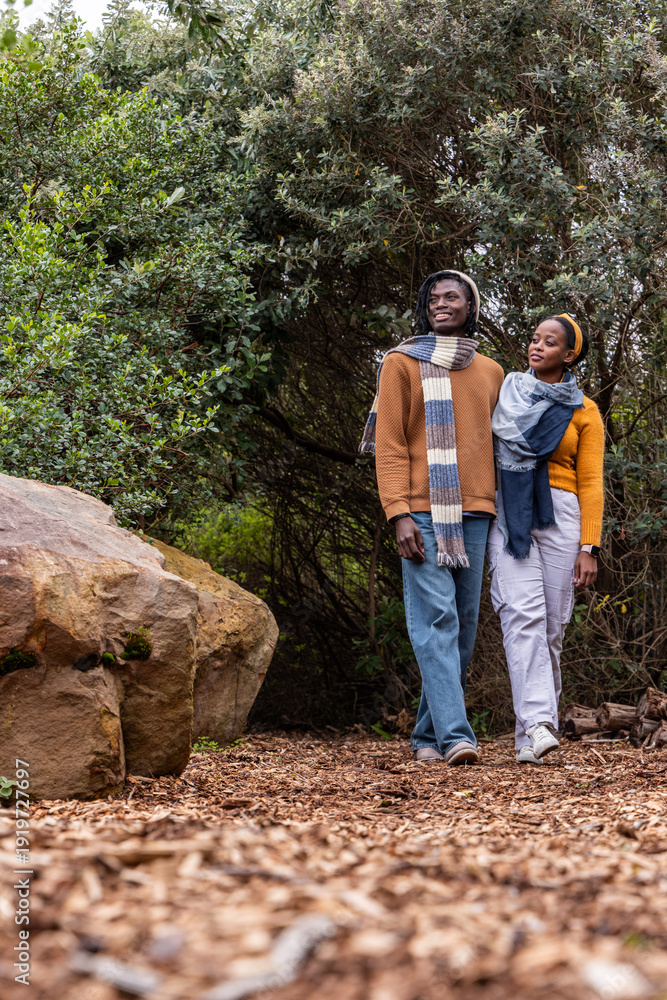 Naklejka premium African American couple walking on woodchip trail near boulder wearing knit scarves and sweaters