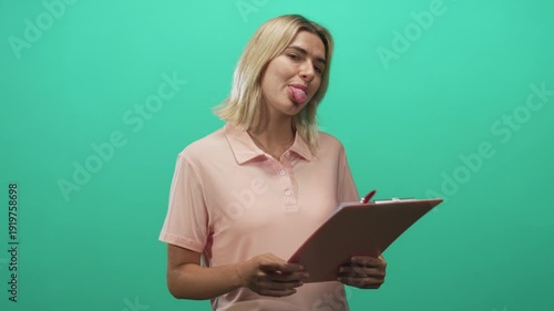 Young blonde hispanic woman holding a pink clipboard sticks tongue out and tilts head toward camera in studio with green backdrop; playful mischief.