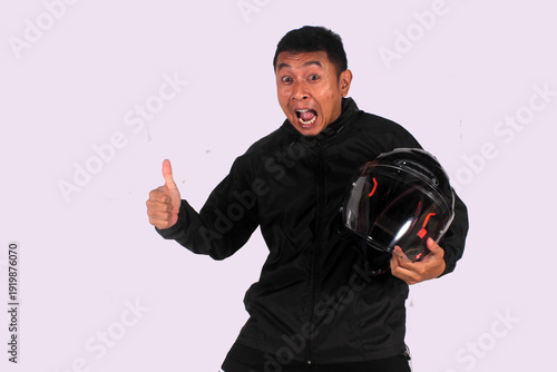 An expressive Indonesian man smiling on an isolated white background while giving a thumbs up (okay symbol) while wearing a jacket carrying a helmet