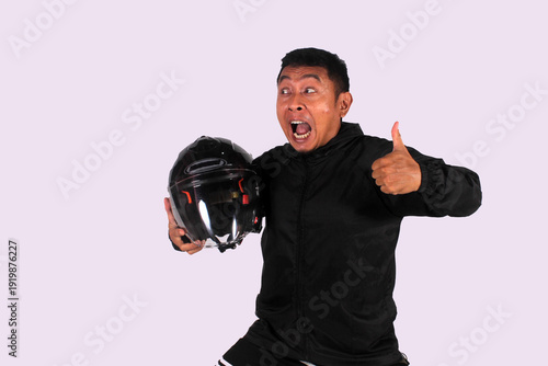 An expressive Indonesian man smiling on an isolated white background while giving a thumbs up (okay symbol) while wearing a jacket carrying a helmet