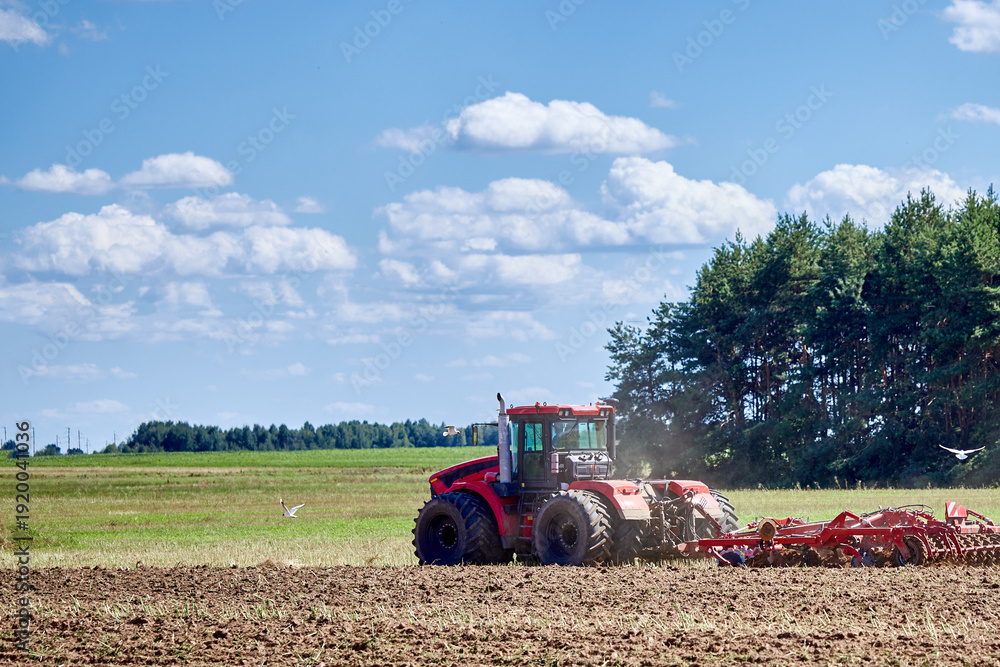 Obraz premium Red tractor cultivating agricultural field, preparing soil for sowing crops, farming industry working in rural landscape
