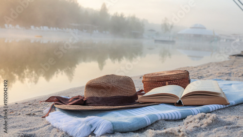 Принти на полотні Relaxing morning by the misty lake with a book and hat on a sandy beach