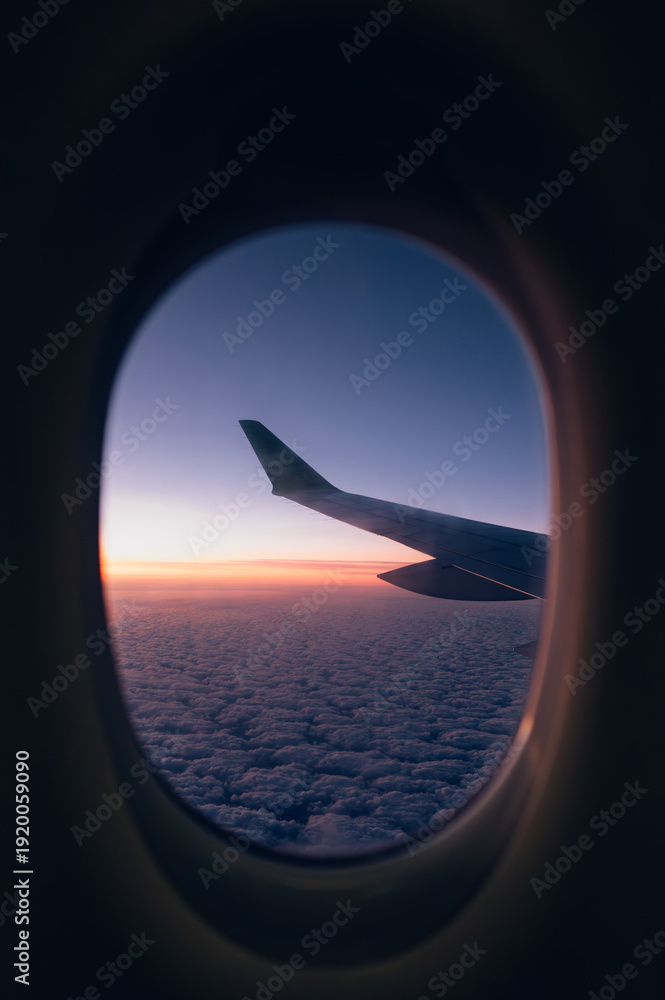 Fototapeta premium Airplane Wing Seen Through Window at Sunrise Above the Clouds