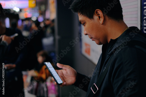 Young Asian man in dark casual wear using smartphone in crowded public space, bokeh blurred background in subway