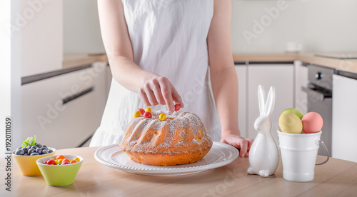 Easter cake being decorated in the kitchen. A person decorating a traditional Easter bundt cake in a bright kitchen setting. The scene includes pastel‑colored eggs, a ceramic bunny, and fresh fruit, c