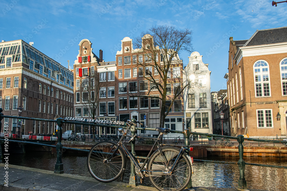 Obraz premium Classic bicycle leans on the seraphina van braziliebrug railing above a tranquil amsterdam canal, framed by gabled houses and historic architecture under a clear blue sky