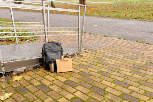 A backpack rests on a brick walkway next to a metal railing with a drink container and a bag
