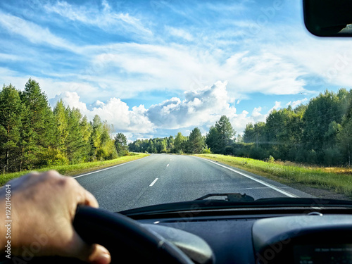 view through car windshield of natural landscape with green forest, white clouds in blue sky. Hand of woman on steering wheel of car. Female traveler