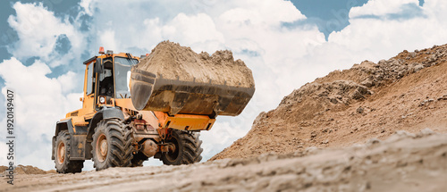 Фотография A powerful excavator loader lifts a heavy load of soil against a backdrop of fluffy clouds