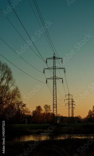 Quiet Landscape With Power Lines And River. Twilight Scene Featuring Pylons Reflecting In Calm River