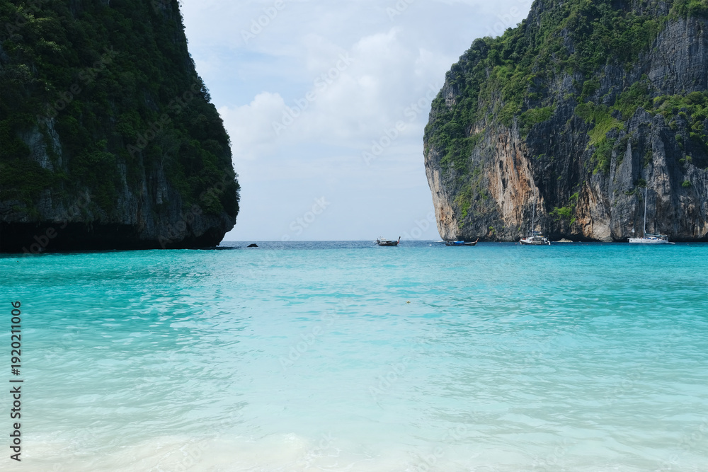 Obraz premium Tropical beach scene with turquoise water, limestone cliffs, and boats anchored near the shore under a partly cloudy sky in a serene coastal environment