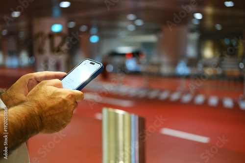 Person using smartphone to call a taxi at an airport terminal with red flooring and blurred background showing vehicle pick-up area