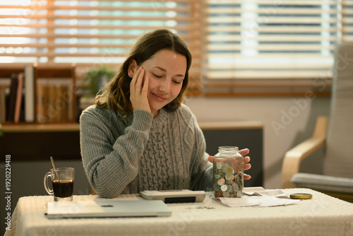 Фотография Young Caucasian woman calculating finances with a jar full of coins, focusing on