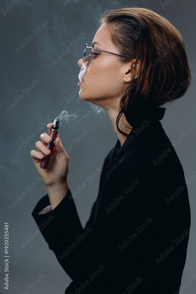 Obraz premium Young woman vaping electronic cigarette with gray background in dark outfit, contemplative mood, modern lifestyle concept.