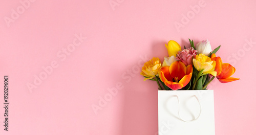 Vibrant bouquet of mixed colorful tulips in white shopping bag on pastel pink background. Spring floral composition with copy space. Greeting card concept for Mother's Day, Women's Day or Easter.