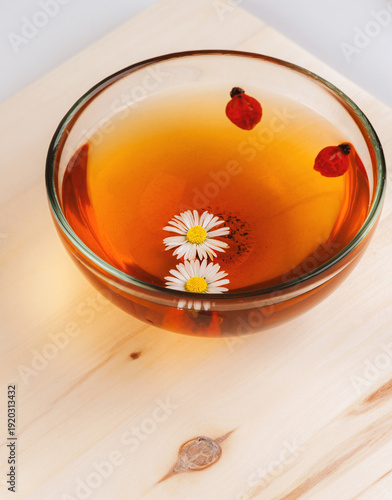 Glass bowl of herbal tea with chamomile flowers and rosehip berries floating in warm amber infusion on wooden table.