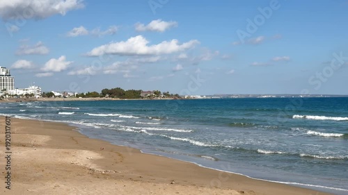 Sunny coastal beach with gentle waves, golden sand, and modern high-rise buildings in the distance. Clear blue sky with fluffy clouds creates a vibrant seaside cityscape and natural ocean beauty
