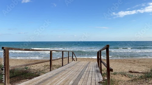 Wooden boardwalk leading to a quiet sandy beach with gentle waves and a blue sea. Sunlight casts creating a serene path to the ocean. Perfect for coastal relaxation and nature-inspired travel concepts