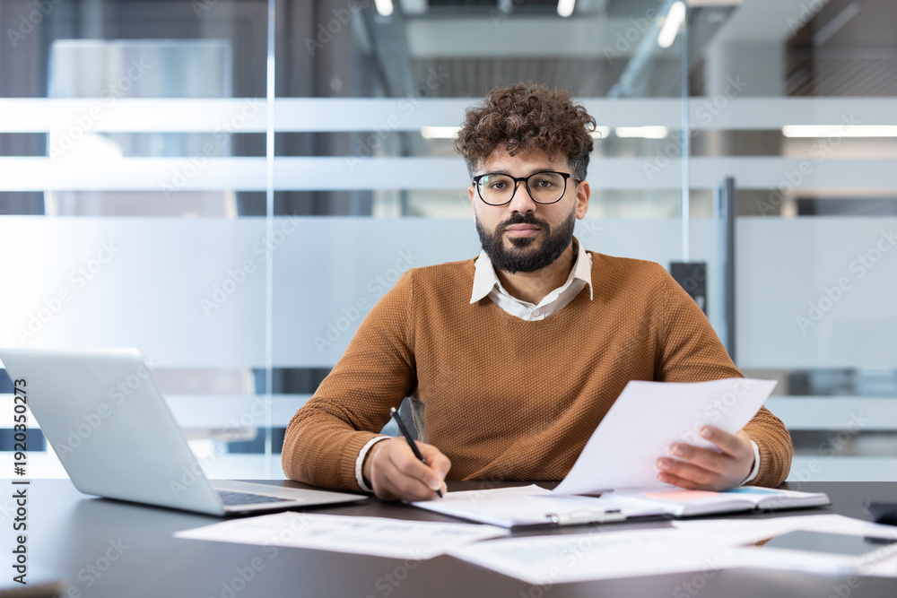 Fototapeta premium Young man with eyeglasses and beard sitting at a desk in a modern office, concentrating on paperwork and using a laptop, representing business analysis and professional work