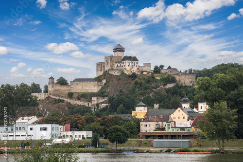 View of medieval Trencín Castle rising above Váh River and colorful houses on green hillside under blue sky with clouds. Iconic Slovak fortress landscape