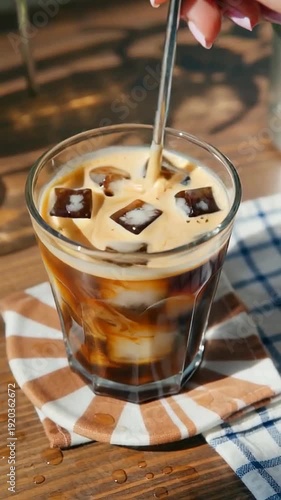 Close-up of iced coffee being stirred with a metal straw in a clear glass, showcasing layers of cream and coffee over ice cubes on a rustic wooden surface
