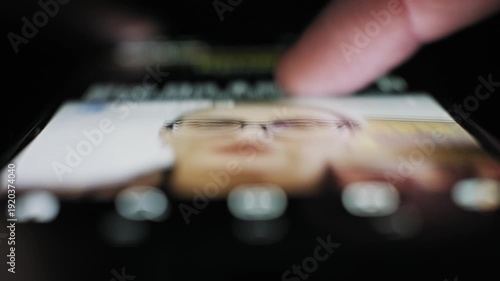 Extreme close-up of a thumb swiping up a touchscreen on a social app news feed in a dark setting. The screen illuminates the finger while scrolling posts photos and video content.