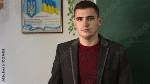 Portrait of a young male teacher in a Ukrainian classroom. Educator standing in front of a chalkboard with Ukrainian flag and coat of arms on the wall