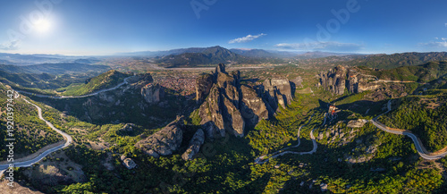 Aerial view of colossal rock formations pierce the verdant landscape, winding roads carving through the terrain below a radiant sun, Kalabak, Trikala, Greece.