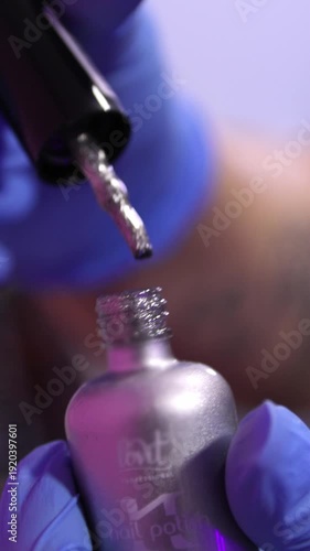 Close-up of a manicurist in blue gloves holding a bottle of silver glitter nail polish. Professional manicure service and beauty salon concept