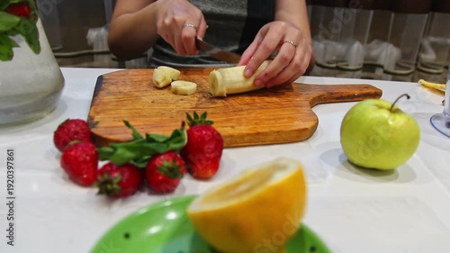 Close-up of a woman slicing a banana on a wooden cutting board. Preparing a fresh fruit salad with strawberries, apples, and oranges. Healthy lifestyle concept