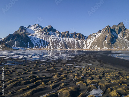 Aerial view of black sand beach contrasting with snow-capped mountains under a clear blue sky, creating a stark but serene landscape, Sveitarfelagid Hornafjordur, Iceland.