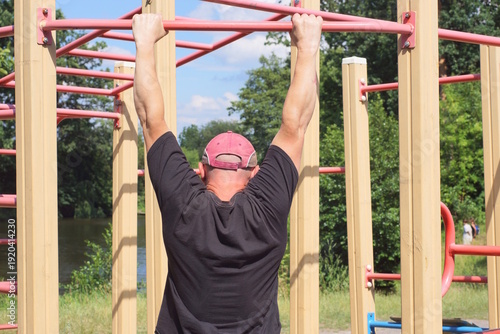 one male athlete in a black shirt pulls himself up on a red sports horizontal bar on the street on a sports ground against a blue sky