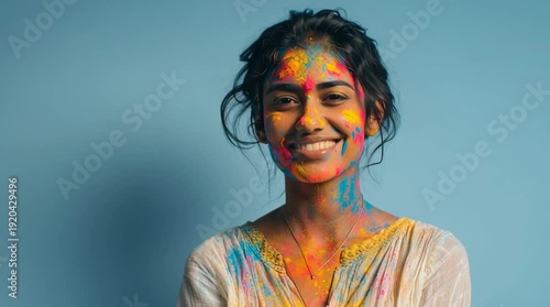 A young South Asian woman smiles with colorful powder on her face. She has dark hair and is wearing a white top against a blue background.