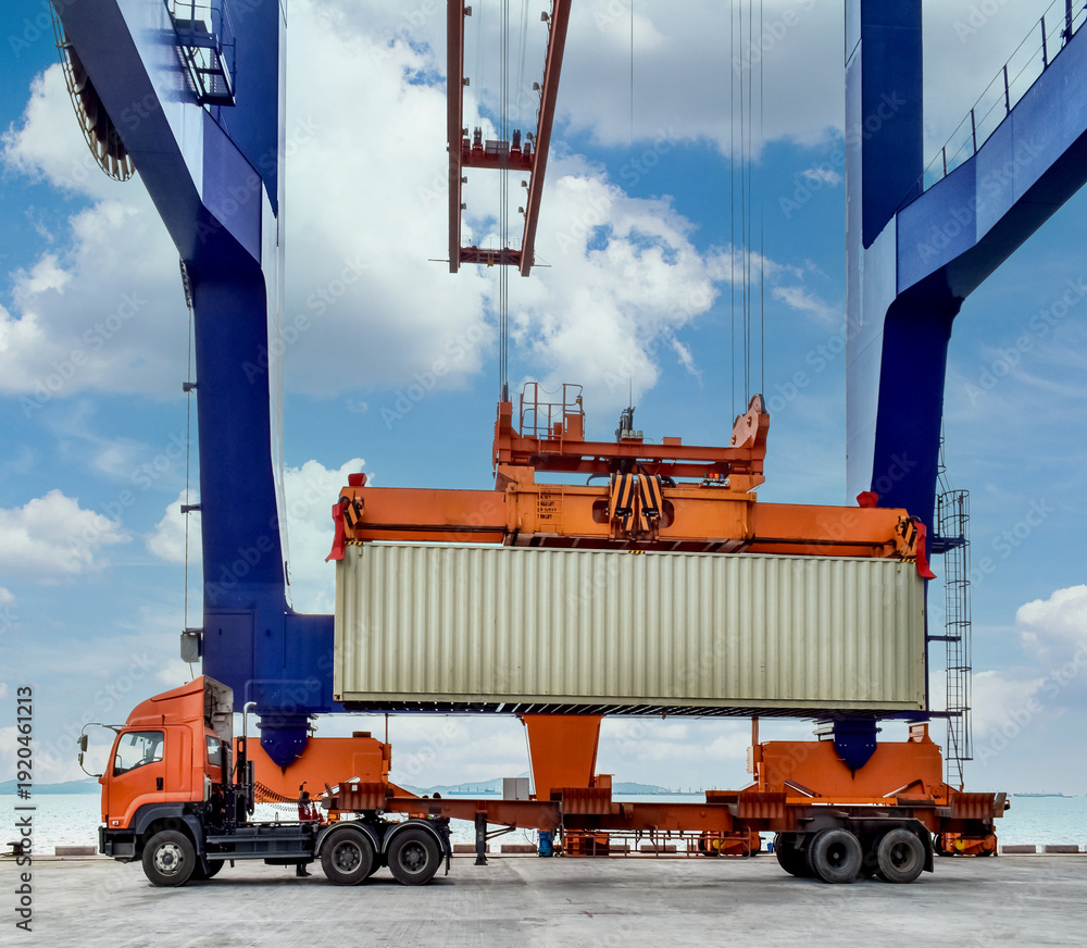 Obraz premium Side view of a massive quay crane loading a shipping container onto a heavy-duty truck at a port terminal.